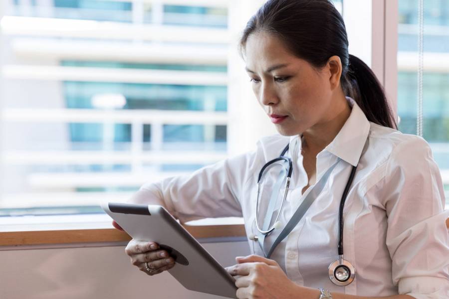 A healthcare provider stands in front of a window with a tablet in her hands.