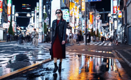 A screenshot from an AI generated clip from Sora AI. A women in a red dress with a leather jacket on top and dark glasses walks confidently down a buys and well lit street which resembles Tokyo. People walk pst in the background and there are many neon signs.