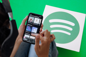 Spotify adds music videos to its streaming platform. A person holds a smartphone displaying the Spotify app with a variety of playlists, against a green backdrop with a large Spotify logo.
