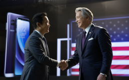 A captivating cinematic scene of a Samsung executive and a suited official from the US government, shaking hands with a smile. The background showcases a sleek Samsung product launch event, with a giant LED screen displaying the latest smartphone and an American flag. The atmosphere is formal yet friendly, with a sense of cooperation and mutual respect., cinematic
