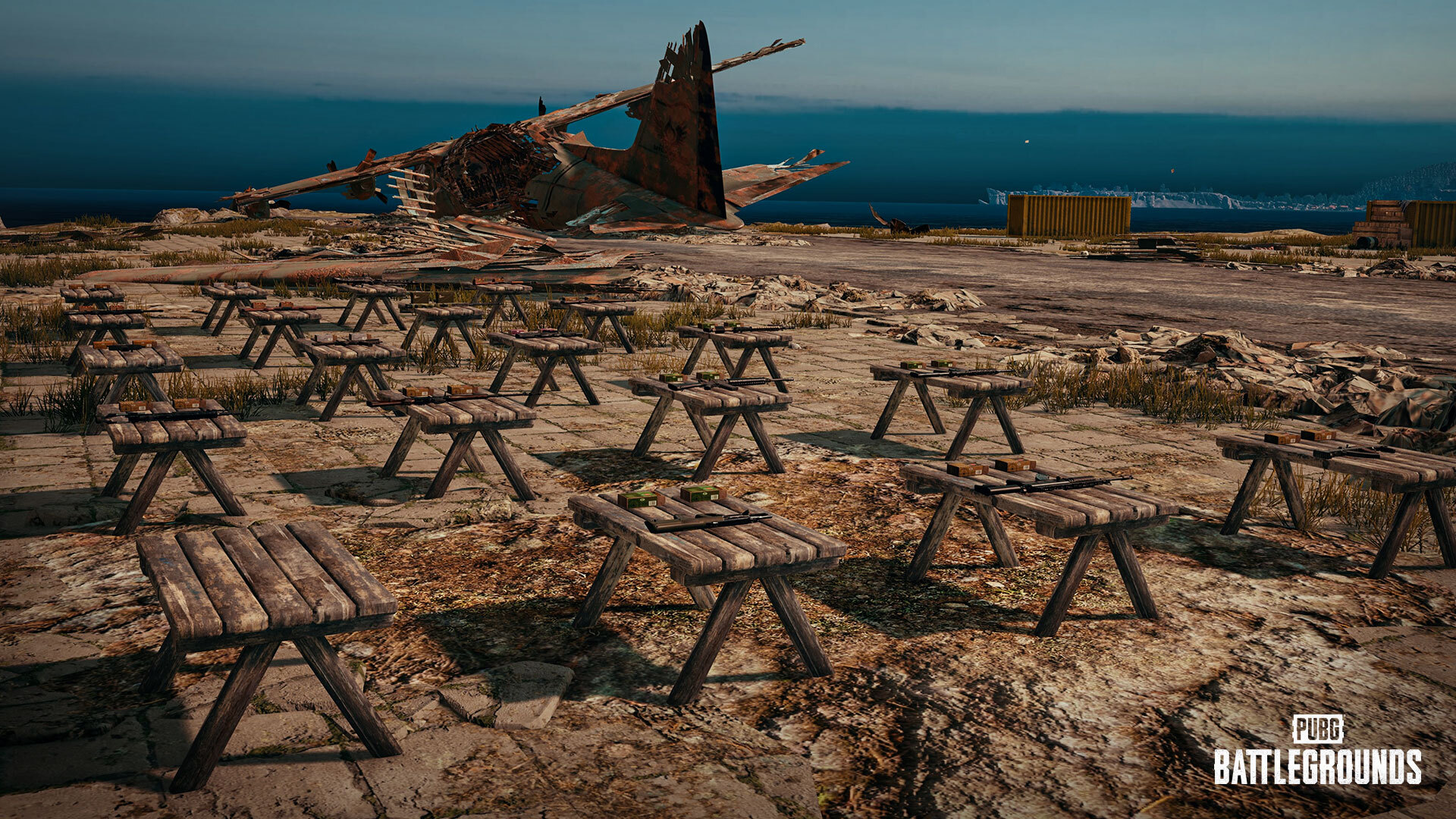 image of weapons arrayed on a grid of picnic benches in PUBG Battlegrounds. A crashed airplane's wreckage is in the distance.