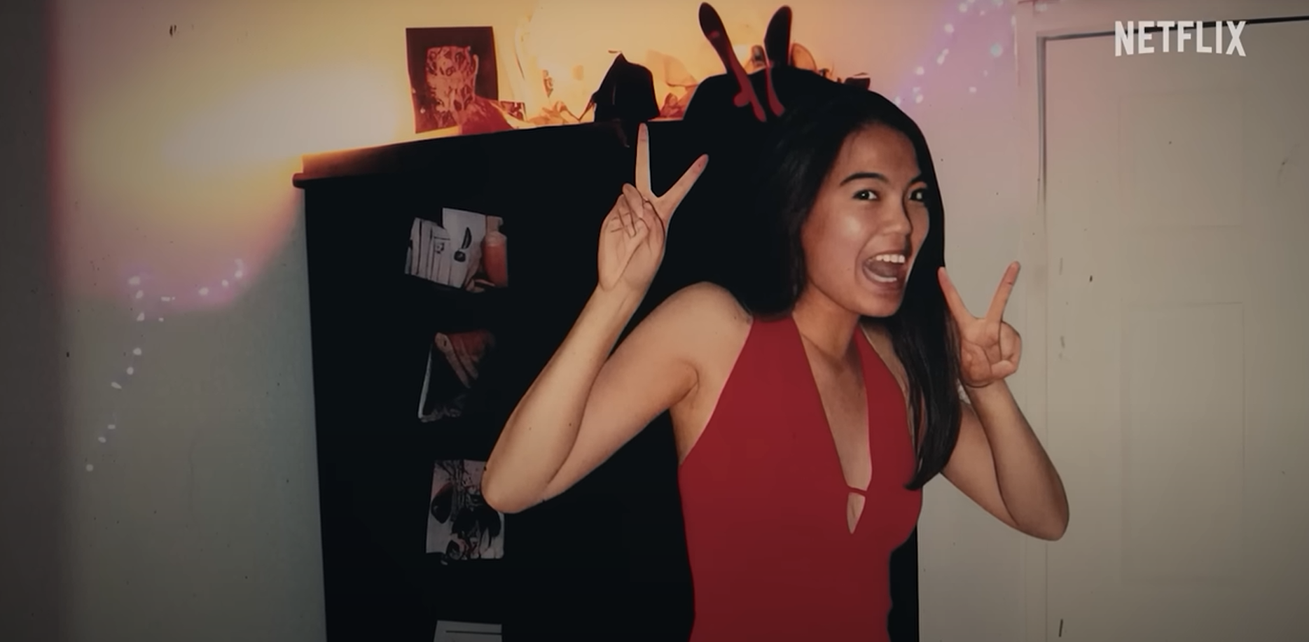 Screenshot of Jennifer Pan from Netflix documentary "What Jennifer Did". A young woman in a red dress stands in a room with a playful expression, making peace signs with both hands. The lighting casts colorful flares across the room, and there are various items and pictures on a dark shelf behind her. "NETFLIX" is displayed in the upper right corner, suggesting this is a screenshot from a Netflix production. A close-up of the same young woman, smiling broadly with her head tilted slightly. She's wearing a red dress, dangling earrings, and has her hand on her shoulder. The background is dark with a few light orbs, indicative of a low-lit environment. "NETFLIX" is visible in the upper right corner, indicating this is also from a Netflix show.