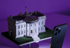 A whimsical miniature replica of the White House, meticulously crafted and placed on a stand. Nearby, a modern smartphone is positioned, seemingly engaged in a video call. The tiny White House is illuminated in purple light, capturing the essence of the iconic building, including its intricate details and green lawn.