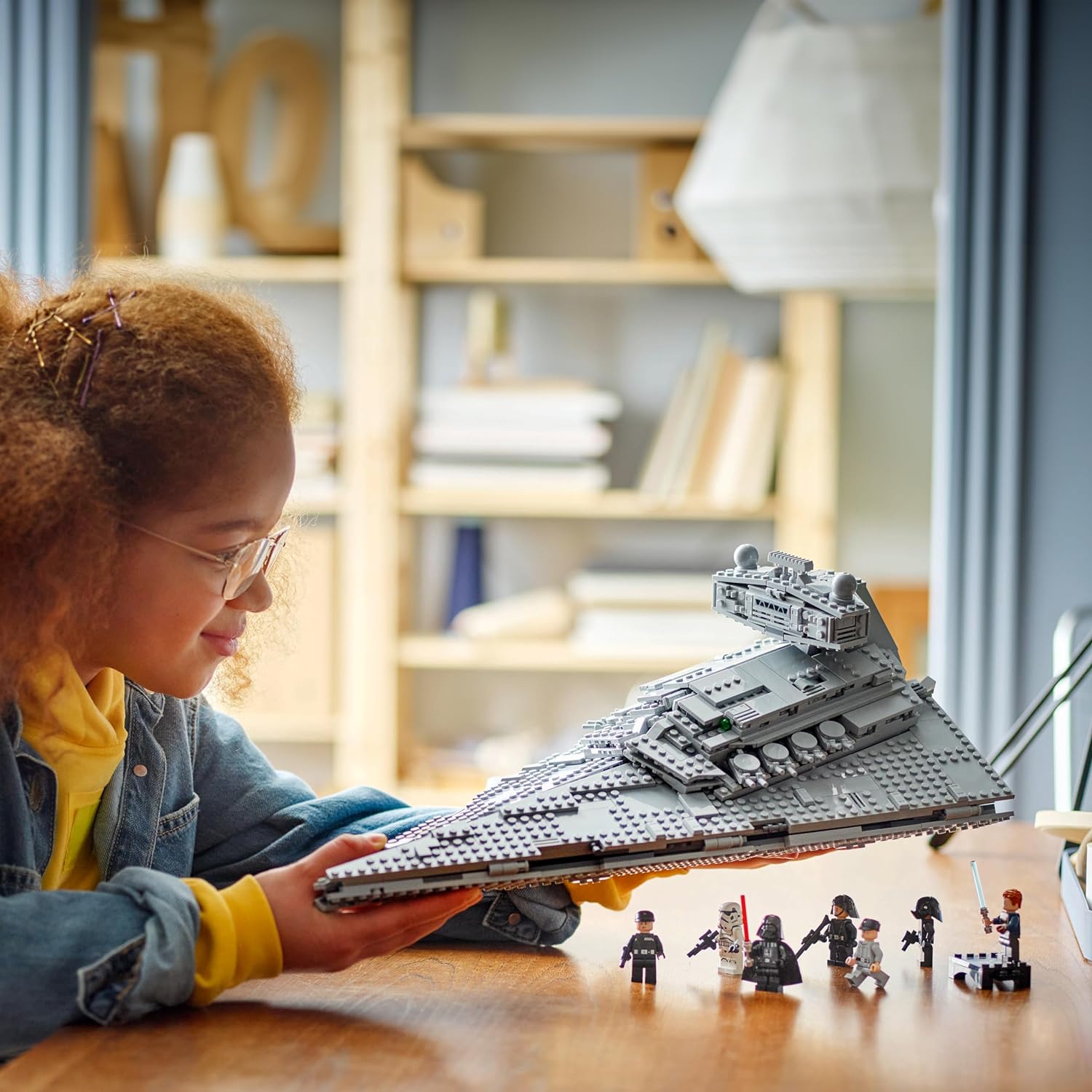A young girl holds up a completed Lego Star Destroyer, showing the iconic Star Wars ship in Lego form.  Seven minifigure characters are on the desk beneath the toy.