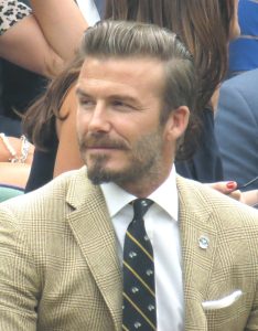 Close up of David Beckham in the Royal Box at Wimbledon in 2014. He's wearing a nude coloured suit with a black tie.