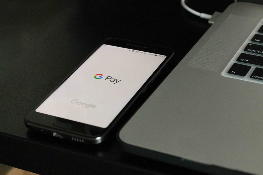 Google Pay on a phone Home Screen on a table. Next to it is a laptop that you can see the left hand side of and some keys