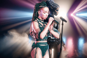 UK MPs call for law to protect musicians from AI deepfakes. An AI-generated image depicting FKA Twigs and an AI-generated version of her on a stage. The primary figure is singing into a microphone, dressed in a futuristic, green and black strappy outfit with braided hair, focused intently on her performance. Beside her, a secondary, more stylized AI figure stands, also with unique ornamented braids and wearing an intricate bodice, under dramatic stage lighting that casts colorful beams and a misty ambiance.