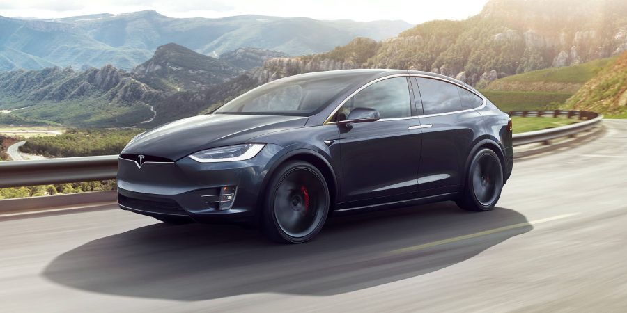 promotional image of a tesla model x driving along a highway against a scenic mountain backdrop