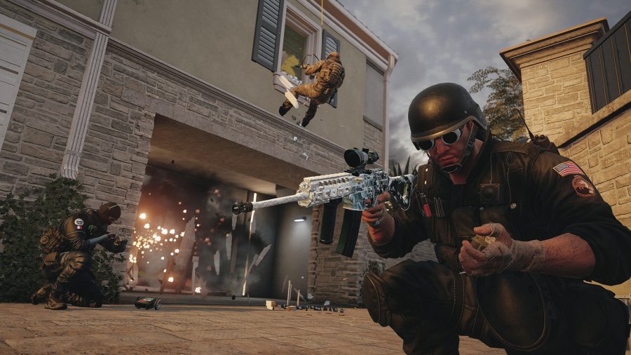 in a Rainbow Six Siege gameplay scene, an operator crouches in the foreground, triggering a detonator that has blown a door open. Another operator in rappelling gear has breached a window and is aiming his weapon through it.