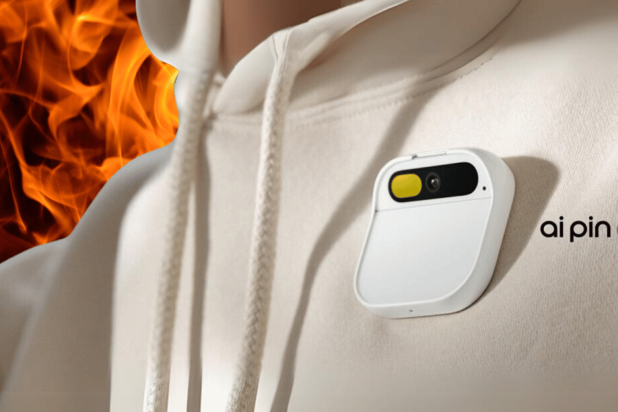 Humane AI Pin charge case poses 'fire safety risk,' users warned. An image showing a small, white charging case for an "ai pin" clipped onto a person's white hoodie. The charging case has a camera lens and a yellow button visible on its front surface. The background features a graphic of orange flames, suggesting a theme of urgency or danger, possibly indicating a fire hazard associated with the product.