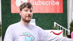 MrBeast in a video wearing a light purple shirt with his logo. Behind him is a dark green bushy background with a red banner displaying the YouTube logo with the text 