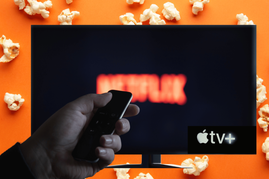Netflix to stop supporting older Apple TV models this summer. A person's hand holding a remote control aimed at a television screen displaying the Netflix logo, with an Apple TV+ logo visible at the bottom right. The background is a vibrant orange, scattered with popcorn, adding a fun, movie-watching ambiance.