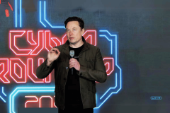 Tesla shareholders approve Elon Musk's $56B pay deal and return to Texas. Elon Musk speaking at an event with a digital background featuring neon-blue and red circuit-like designs. He is dressed in a casual jacket, holding a microphone, and gesturing with his hand as he speaks. The setting suggests a high-tech or futuristic theme, aligning with the technological focus of the event.