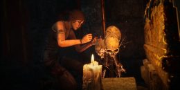 Lara Croft rigs up what looks like some kind of booby trap using a collection of human skulls. The room is dimly lit by candle and Croft is flecked with grime, in her signature "Survivor Croft" look.