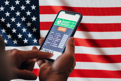 Flutter boosts US presence while shifting away from UK and strict gambling laws. A person holding a smartphone displaying a sports betting app screen, with the American flag in the background.