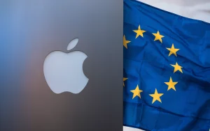 Uma foto de um logotipo da Apple com uma bandeira da UE ao lado. O logotipo da Apple está em um fundo cinza, e a bandeira da UE está em um fundo branco. A bandeira da UE tem 12 estrelas amarelas em um fundo azul. A imagem tem uma iluminação suave.