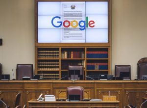 A photo of a European court with the Google logo on a screen. The screen is displaying a legal document. The room has wooden furniture, including a desk and chairs. There are books on shelves behind the desk. The walls are painted beige.