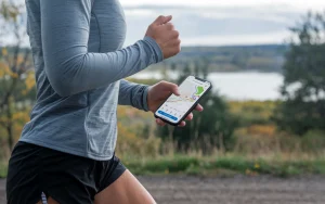 An image of a person wearing a gray shirt and black shorts, going for a run outdoors. The person is holding a smartphone with the Strava app open, showing a map of the run route. The background is a scenic landscape with trees and a body of water.