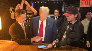 Donald Trump surrounded by people at PubKey bar in NYC. He's stood at the bar where he bought burgers using Bitcoin. He's on his phone.