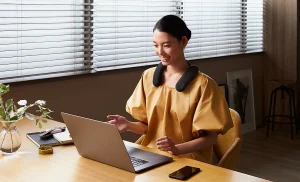 Woman sat at a desk wearing the Sony Bravia Theatre U Neckband. She's on her laptop taking a call in a dimly lit office.