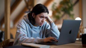 A cinematic shot of a person sitting at a table, using a MacBook. The person is wearing a gray hoodie and has long, dark hair. They are looking frustrated because the MacBook's screen is black. The laptop is placed on a wooden surface. There are a few objects on the table, including a mug and a notebook. The background is blurred, revealing a cozy room with wooden beams and a plant.