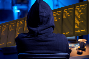 Police investigate cyberattack on UK train station WiFi network. Hooded hacker in front of train display screens