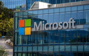 A photo of a building with the Microsoft logo. The building has multiple floors and is made of glass. The Microsoft logo is prominently displayed on the side of the building. The background contains trees and a road.