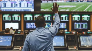 A photo of a man placing a sports bet at a betting window in a New York City sportsbook. The man is wearing a blue shirt and has his hand raised. The background contains multiple betting windows and a large screen displaying a sports event.