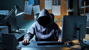 A photo of a shadowy hacker sitting in a dimly lit room. The hacker is wearing a hoodie and has their face obscured. They are sitting at a computer desk. The room has various electronic devices, including a monitor, keyboard, mouse, and a lamp. The wall behind the hacker has a bulletin board with papers pinned to it.