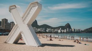 A photo of Copacabana Beach in Rio de Janeiro, Brazil with a massive neon white "X". The "X" is located on a building in the background and casts a shadow on the sand. The beach is crowded with people relaxing and playing in the sand. The water is calm and has a few people swimming. The sky is clear with a few clouds.