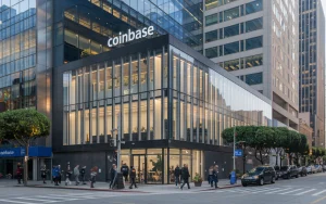 A photo of the exterior of Coinbase's headquarters in San Francisco. The building has a modern design with a glass façade. The building is located in a bustling area with other buildings nearby. There are people walking on the sidewalk. The building has a Coinbase sign on the top floor. The background contains tall buildings. The overall image has natural lighting.