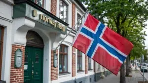 A photo of a bank building in Norway with the Norway flag next to it. The bank has a traditional architecture with red and white bricks and a green door. The Norway flag is waving in the wind. The background contains trees and a street.