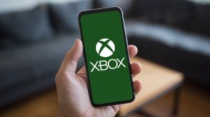 A photo of a mobile phone with the Xbox logo on the screen. The phone is in a hand. The background is blurred and contains a couch and a table.