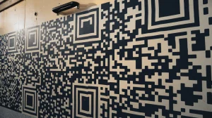 A photo of a wall covered in large QR codes. The QR codes are arranged in a pattern, with some overlapping. The background is a beige wall with a few dents. There's a black handle on the wall near the bottom. The lighting is bright.