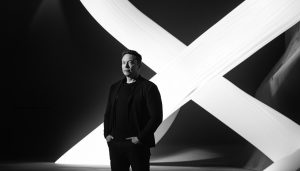 Black and white Elon Musk standing in front of a large white 'X' on a dynamic background