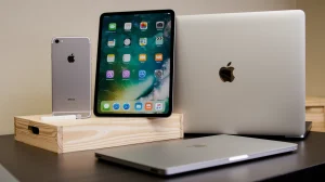 A photo of a table with several Apple devices. There is an iPhone, an iPad, and a MacBook Pro. The iPhone and iPad are placed on a wooden box, and the MacBook Pro is placed next to the box. The devices are placed in the foreground, against a beige wall in the background.