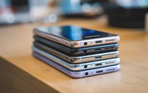 A photo of a stack of Samsung phones. The phones are placed on a wooden surface. The topmost phone is slightly tilted, revealing the Samsung logo. The background is blurred and contains a few other objects.