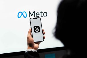 White screen with the Meta logo on in the background, with person holding a phone with the Threads app on it.