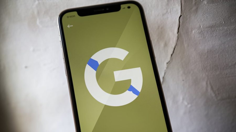 A photo of an iPhone displaying a Google logo. The Google logo is a large, white "G" on a green background, centered on the screen. The background is a white wall, with a few cracks. The lighting is soft.