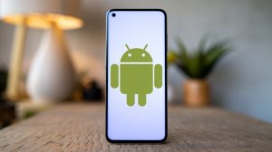 A photo of a phone with the Android logo displayed prominently on the screen. The phone is placed on a wooden surface. The background is blurred and contains a few objects, including a lamp and a plant. The lighting is soft.