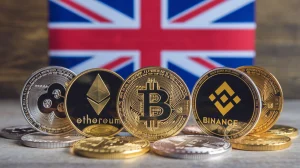 A photo of multiple crypto coins, including Bitcoin, Ethereum, and Binance Coin. There is a UK flag in the background. The coins are placed on a wooden surface. The background is blurred.