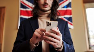 A photo of a person holding a smartphone, using social media. The person has long hair and is wearing a beige turtleneck sweater and a navy blue blazer. The background contains a UK flag hanging on the wall. The room has a beige wall, a wooden door, and a window. The lighting is warm.