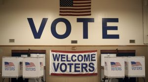 A photo of a US polling station with the word 'VOTE' written on the wall. There are voting booths inside the station. The station has a flag and a banner with the words 'Welcome Voters'. The overall ambiance is serious and peaceful.