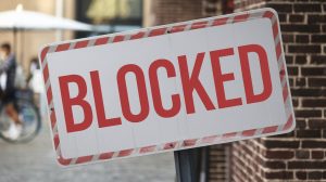 A photo of a large sign with the text 'BLOCKED' in bold, red letters. The sign is placed against a brick wall. The background is blurred and contains a few people and a bicycle. The lighting is soft.