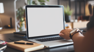 A photo of a laptop with a blank screen. The laptop is on a desk with a few objects such as a pen, a notebook, and a phone. A person's hand with a watch is visible, holding the laptop, with a bent wrist. The background is blurred and contains a plant, a lamp, and a wall.