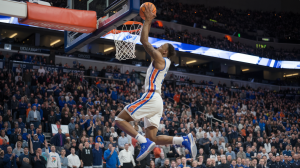 A basketball game with a packed stadium. A player in a white jersey with blue and orange stripes is in mid-air, attempting a slam dunk. The basket is orange and has a white net. The background is filled with cheering fans.