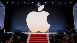 A photo of a large Apple logo with a trophy on top of it. The logo is placed on a stage at an awards show. The stage has a red carpet leading up to it. There are multiple cameras pointed at the logo. The background is dark.