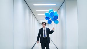 Adam Scott actor in a white sterile hallway, dressed in a suit. He's holding a bunch of light and dark blue balloons with his face on them.