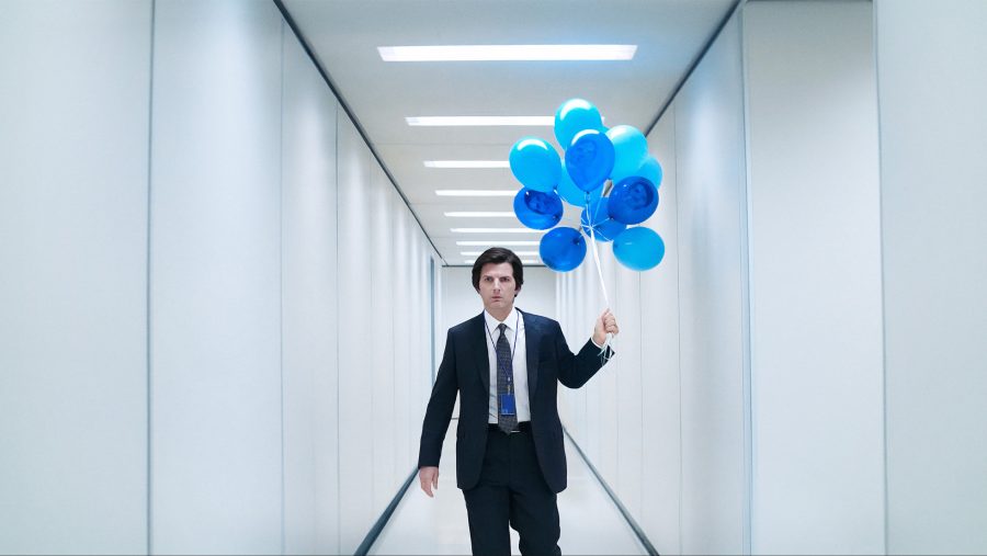 Adam Scott actor in a white sterile hallway, dressed in a suit. He's holding a bunch of light and dark blue balloons with his face on them.