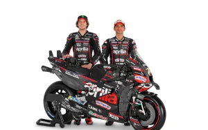 Two men stood in race gear behind a Moto GP Aprilia Racing bike.